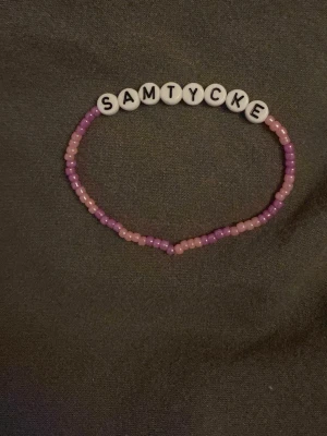 Pärlarmband med texten SAMTYCKE - Ett handgjort armband med små rosa och lila pärlor samt vita bokstavspärlor som stavar 'SAMTYCKE'. Armbandet har en enkel och lekfull design, perfekt för att uttrycka din personlighet och budskap. Trädd på elastisk tråd för enkel användning.