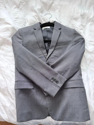 Grå kavaj från H&M - Stilren grå kavaj från H&M med två knappar framtill, klassisk krage och två fickor med lock. Kavajen har en bröstficka och är helfodrad med svart innerfoder. Perfekt för en dressad look.