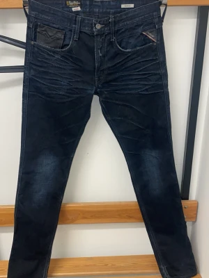 Svarta Replay jeans - Snygga svarta jeans från Replay med slitna detaljer och markerade sömmar. Jeansen har klassisk femficksdesign, smal passform och coola kontrastsömmar vid fickorna. Perfekta för en trendig och avslappnad stil.