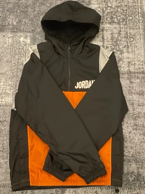 Jordan vindjacka svart orange - Snygg vindjacka från Jordan i svart och orange med vita detaljer på axlarna. Jackan har huva, dragkedja framtill och elastiska muddar. Stor logga på bröstet. Perfekt för streetwear och sportiga outfits.