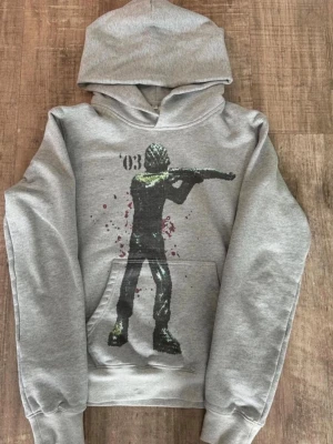 Grå hoodie med tryck av skjutare - Snygg grå hoodie med en stor tryckt figur på framsidan som håller ett gevär. Hoodien har huva och en klassisk magficka. Perfekt för dig som gillar streetwear och vill sticka ut med ett unikt motiv.