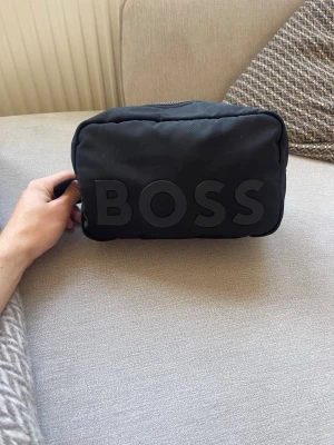 Hugo Boss necessär – stilren svart – perfekt för resor - Stilren Hugo Boss necessär i svart. Perfekt storlek för resor eller gym. Diskret BOSS-logo och kraftig dragkedja.  • Märke: Hugo Boss • Färg: Svart • Skick: Mycket bra • Rymlig och praktisk • Perfekt till resor / gym / vardag  Säljes pga garderobsrensning. Skickas samma dag