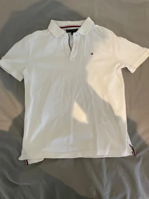 Vit pikétröja Tommy Hilfiger - Vit pikétröja från Tommy Hilfiger med klassisk krage, knappar framtill och liten broderad logga på bröstet. Tillverkad i bomull och har korta ärmar samt diskreta rödblå detaljer på insidan av kragen och nederkanten. Storlek 158-164. Kragen är lite fläckig men märkts knappt. 