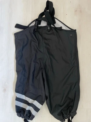 Svarta täckbyxor med hängslen - Svarta  galonbyxor  med justerbara hängslen och reflexdetaljer på benen. Byxorna har resår vid bensluten och knappar upptill. Perfekta för regniga dagar och utomhusaktiviteter. Materialet är slitstarkt och vattenavvisande.