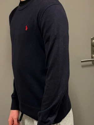 Mörkblå tröja från Polo Ralph Lauren - Pris kan diskuteras!   Killen i bilden är 172 cm lång, den är lite kort på han. Så jag skulle föreslå att man ska vara runt 160-170.        Mörkblå långärmad tröja från Polo Ralph Lauren med klassisk rund hals och röd broderad logga på bröstet. Tröjan är stickad och har ribbade muddar vid ärmslut och nederkant. Perfekt för en stilren och avslappnad look.