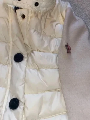 Vit dunväst från Moncler - Snygg vit dunväst från Moncler med svarta knappar och broderad logga på ärmen. Jackans design och Materialet är dunfyllt och ger en lyxig känsla.