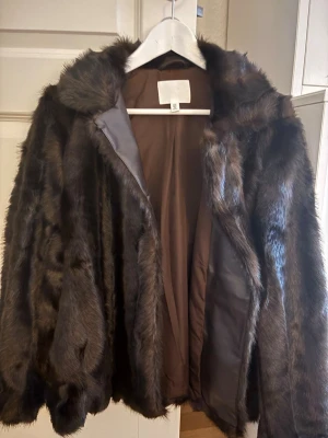 Mörkbrun pälsjacka med krage - Snygg mörkbrun pälsjacka med klassisk krage och helbrun insida. Jackan har en rak passform och är tillverkad i mjukt fuskpälsmaterial som ger en lyxig känsla. Perfekt för att hålla värmen under kalla vinterdagar.