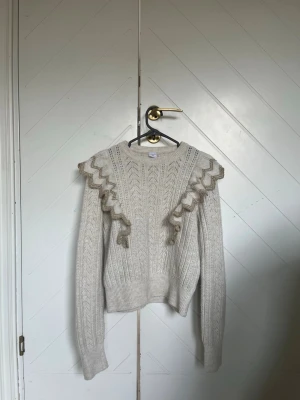 Beige stickad tröja med volang - Snygg beige stickad tröja med dekorativa volanger längs axlarna och ett fint mönster i stickningen. Tröjan har lång ärm, rund halsringning och ribbade muddar. Perfekt för dig som gillar en trendig och mysig look.😍❤️