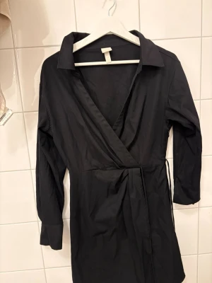 Svart omlottklänning från H&M - Svart omlottklänning från H&M med lång ärm och klassisk krage. Klänningen har knyt i midjan och en stilren, enkel design som passar perfekt till många tillfällen. Tillverkad i ett mjukt och lätt material för en bekväm känsla.