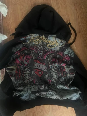 Ed Hardy hoodie med tryck - Säljer en svart Ed Hardy hoodie med stort färgstarkt tryck på ryggen, dekorerad med guldfärgade nitar. Hoodien har dragkedja där fram. Lagt till det vita way out west trycket. Hoodien är sliten i tröjärmarna.