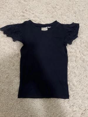 Svart topp med volangärmar - Svart topp med rund halsringning och korta volangärmar i broderad spets. Toppens kropp är i mjuk, stretchig bomullskvalitet och ärmarna har fina håldetaljer som ger en söt och trendig look.