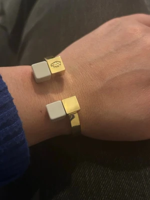 Iaoku armband med vita kuber - Snyggt armband i metall med guldfinish och två vita kubformade detaljer i ändarna. Ena kuben har en liten ingraverad symbol som ger en unik touch. Armbandet har en modern och stilren design som sticker ut.