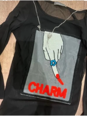 Svart meshtröja med CHARM-tryck - Cool svart meshtröja från UNIF med ett stort tryck framtill som visar en hand med röd nagellack och blå ring, samt texten CHARM i rött. Tröjan har lång ärm och transparent material som ger en edgy vibe.