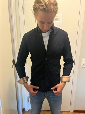 Marinblå burberry skjorta - Säljer en svart skjorta med klassisk krage och knappar framtill. Manschetterna har ett rutigt mönster i beige, rött och svart som ger en snygg kontrast. Skjortan är långärmad och har en normal passform, perfekt för dig som vill ha en stilren look.