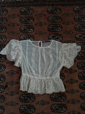 Vit spetsblus med volang  - Vit spetsblus från Zara med transparent mesh och ett broderat blommönster. Blusen har korta, puffiga volangärmar och rund halsringning med knapp i nacken. Midjan är markerad med volangkant. Nästintill oanvänd.