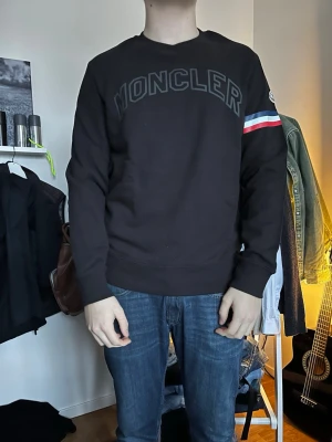 Svart sweatshirt från Moncler - Säljer en svart sweatshirt från Moncler med stor logga på bröstet och en snygg detalj med röd, vit och blå rand på ena ärmen. Tröjan har rund halsringning och är tillverkad i mjukt material, perfekt för en avslappnad och stilren look.