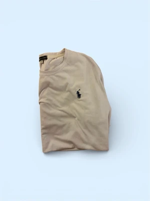 Vit t-shirt från Ralph Lauren - Klassisk beige t-shirt från Ralph Lauren med den ikoniska mörkblå polospelaren broderad på bröstet. T-shirten har rund hals och korta ärmar, perfekt för en clean och stilren look. Står L men passar som m