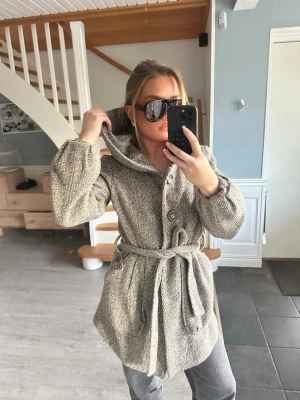 Beige ullkappa med knytskärp - Snygg beige kappa i ullmix med bred krage, stora knappar och knytskärp i midjan. Kappan har två stora fickor och en avslappnad passform som ger en cool look. Perfekt för kalla dagar och passar till jeans eller dressade byxor.