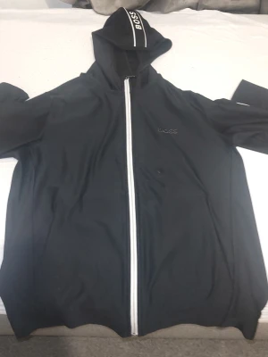 Svart hoodie med dragkedja från BOSS - Svart hoodie från BOSS med vit dragkedja och logga på bröstet, huvan och längs ärmarna. Snygg sportig look med vita detaljer och huva. Perfekt för dig som gillar streetwear och vill ha något stilrent med tydlig logga.