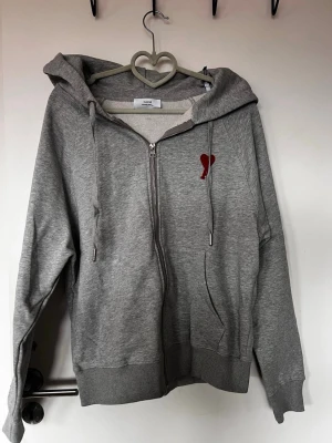Grå hoodie med dragkedja från Ami - Snygg grå hoodie från Ami med dragkedja och huva. På bröstet finns ett rött broderat hjärta med A. Hoodien har ribbade muddar och fickor på sidorna. Tillverkad i mjuk bomullsblandning, perfekt för en chill och stilren look.