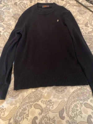 Mörkblå stickad tröja från Morris - Snygg mörkblå stickad tröja från Morris med rund halsringning och diskret logga broderad på bröstet. Tröjan har ribbade muddar vid ärmslut och nederkant, vilket ger en klassisk och stilren look. Perfekt för dig som gillar enkel men elegant stil.