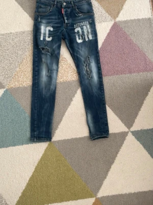 Blå Dsquared2 ICON jeans - Snygga blå jeans från Dsquared2 med vit 'ICON'-text framtill. Jeansen har slitna detaljer och hål på båda benen för en trendig look. Klassisk femficksmodell med smal passform och knappgylf.