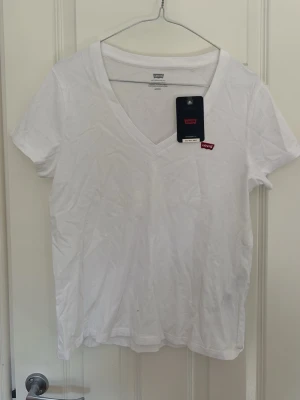 Vit v-ringad t-shirt från Levi's - Vit t-shirt från Levi's med v-ringning och klassisk röd logotyp på bröstet. Standard fit och tillverkad i mjuk bomull, perfekt för en clean och enkel stil. Passar till allt och är superbekväm att bära hela dagen.