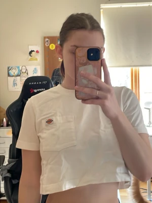 Vit croppad t-shirt från Dickies - Säljer en vit croppad t-shirt från Dickies med korta ärmar och bröstficka med logotyp. Perfekt för dig som gillar streetwear och vill ha en enkel men snygg look. Materialet är bomull och modellen är normal passning.