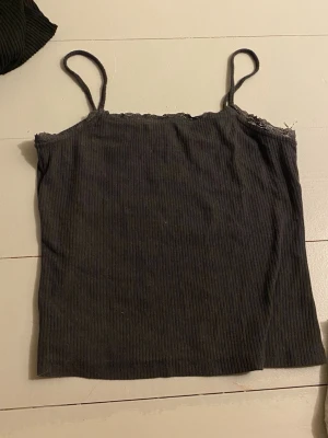 Svart ribbad linnetopp med spetskant - Säljer en svart ribbad linnetopp med tunna axelband och en diskret spetskant upptill. Perfekt att styla med jeans eller kjol för en enkel och snygg look. Materialet känns mjukt och följsamt.