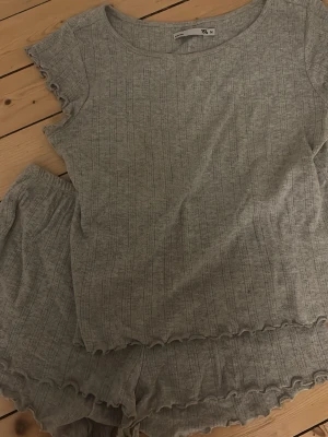 Grå ribbad pyjamas-set - Mysigt pyjamas-set i ljusgrått ribbat material. Setet består av en kortärmad topp med rund hals och volangkant samt matchande shorts med volangdetaljer. Perfekt för chillkvällar hemma.