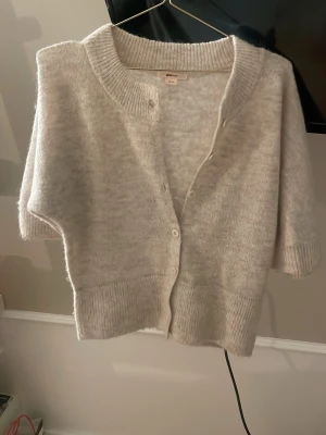 Ljusgrå kofta med kort ärm - Mysig ljusgrå kofta med trekvartsärmar och knappar framtill. Koftan har bred ribbstickad mudd vid ärmslut och nederkant samt en djup v-ringning. Perfekt för lager-på-lager och enkel att matcha.