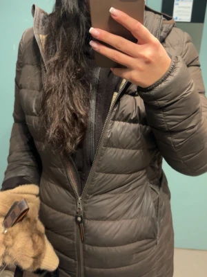 Brun lång dunjacka från Parajumpers - Snygg brun dunjacka från Parajumpers med huva och dragkedja hela vägen. Jackan är quiltad och har en längre modell som håller dig varm under kalla dagar. Den varmaste jackan som jag ägt, har hållt mig varmare än andra vinter vinter jackor. Märkeslogga på ärmen och praktiska fickor på sidorna. Det finns 2 rispor på bröst och arm som säkert går att laga men har inte testat själv. 