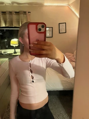 Ljusrosa croppad topp med knyt - Trendig ljusrosa croppad topp med långärmad design och knyt framtill. Toppen har en ribbad struktur och flera öppna detaljer längs bröstet som ger en cool look. Perfekt för dig som gillar att sticka ut och vill ha något unikt i garderoben.