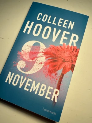 9 November av Colleen Hoover - En gripande roman av Colleen Hoover med blått omslag, stora vita bokstäver och en röd blomma. Perfekt för dig som gillar romantik och starka känslor. Boken är på svenska och har ett mjukt omslag.