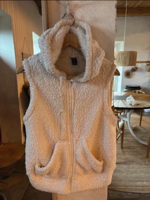 Vit teddyväst från Emery Rose - Mysig vit teddyväst från Emery Rose med dragkedja framtill och två stora fickor. Västen har huva och är tillverkad i fluffigt teddy-material som ger en riktigt chill vibe. Perfekt att slänga över en hoodie eller långärmad tröja för extra värme. Hoodie liknande