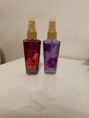 Victoria's Secret Pure Seduction & Love Spell - Två body mists från Victoria's Secret: Pure Seduction i rödrosa flaska med blommigt motiv och Love Spell i lila flaska med blommigt motiv. Båda har guldfärgade spraymunstycken och rymmer 60 ml. Doftnoter av röda plommon, fresia, körsbärsblom och persika.