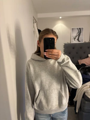 Grå hoodie från Gina  - En klassisk ljusgrå hoodie med huva och stor magficka framtill. Tröjan har ribbade muddar vid ärmslut och nederkant, och är tillverkad i mjukt sweatshirt-material. Perfekt för en avslappnad och cool look.
