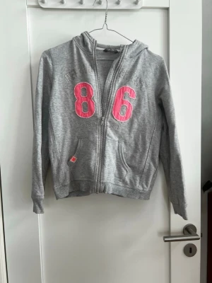 Grå hoodie med rosa 86-tryck - Snygg grå hoodie med dragkedja från H&M. Stor rosa 86-tryck framtill, två fickor och huva. Perfekt för en avslappnad och sportig stil. Mjuk bomullsblandning som är skön att ha på sig hela dagen. Storlek 146-152 så typ xxs🫶🏽 en knapps synlig fläck se sista bilden