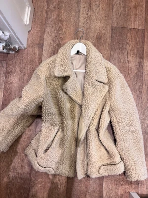 Beige teddyjacka med dragkedjor - Säljer en beige teddyjacka med bred krage och två snedställda dragkedjefickor. Jackan har en mysig, fluffig yta och är fodrad på insidan. Perfekt för kalla dagar och ger en trendig look till din outfit.