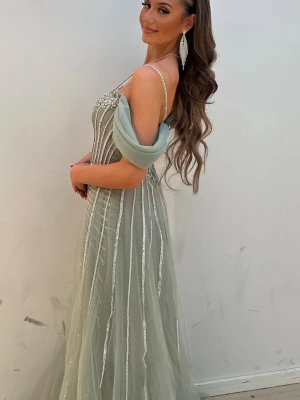 Glittrig långklänning med silverdetaljer - Elegant långklänning i ljus grön ton med glittriga silverränder och offshoulder-design. Klänningen har ett vackert fallande tyg och dekorativa detaljer vid axlarna. Perfekt för speciella tillfällen och bal.