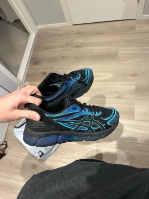 Svarta och blå Asics sneakers - Säljer ett par svarta Asics sneakers med blå detaljer och mönster längs sidorna. Skorna har en chunky sula i svart och blått och är låga i modellen. Perfekta för dig som vill sticka ut med en sportig och modern look.