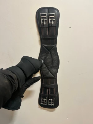 Horka sadelgjord 50cm - Säljer en svart sadelgjord från Equor i mjukt läder med ergonomisk form och rejäla spännen i metall. Gjorden har elastiska partier och en D-ring i mitten för enkel fastsättning av tillbehör. Perfekt till hästutrustning.