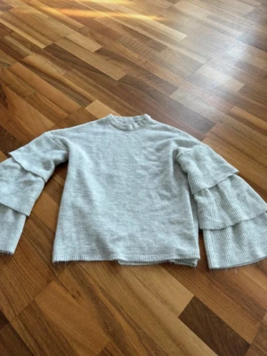 Ljusgrå stickad tröja från ONLY - Trendig ljusgrå stickad tröja från ONLY med volangärmar i två lager och rund halsringning. Mjuk och skön kvalitet, perfekt för dig som vill ha en unik detalj på din stickade tröja. Passar till både jeans och kjol.