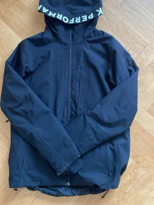 Mörkblå vindjacka Peak Performance - Snygg mörkblå vindjacka från Peak Performance med huva och dragkedja framtill. Jackan har logga på huvan och är tillverkad i ett slitstarkt, vattenavvisande material. Perfekt för blåsiga dagar och outdoor-aktiviteter.