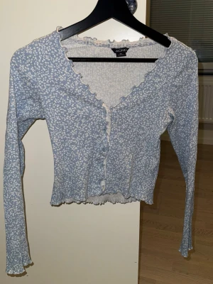 Blommig blå kofta från Monki - Supersöt blå kofta från Monki med små vita blommor, volangkant längs v-ringningen och knäppning framtill. Långärmad och croppad modell i mjukt, stretchigt material. Perfekt för en trendig och avslappnad look.