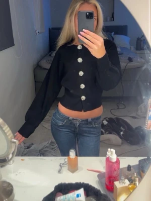 Svart kofta med glittriga knappar - Snygg svart kofta med långa ärmar och stora glittriga knappar framtill. Kortare modell som slutar vid midjan och har en enkel, stilren design. Perfekt för att lyfta en outfit med lite extra detaljer.