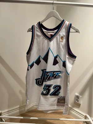 Utah Jazz | Karl Malone #32 - Snyggt vitt basketlinne från Mitchell & Ness med Utah Jazz-logga och nummer 32. Klassisk design med bergsmotiv, lila och turkosa detaljer samt namnet Malone på ryggen. Tillverkat i ventilerande meshmaterial, perfekt för basket eller streetwear. Ihoplimmad  baktext och inte så flitigt använd. 8,5/10