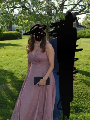 Glittrig rosa långklänning omlott - Säljer en glittrig rosa långklänning med omlottliv och v-ringning. Klänningen är ärmlös och har ett elegant fall, perfekt för speciella tillfällen. Materialet skimrar snyggt i ljuset och ger en lyxig känsla. Använd en gång. Storlek L men passar även mig med M