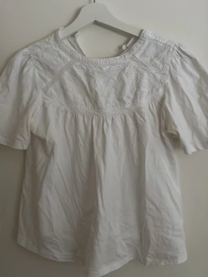 Vit blus med broderi och puffärm - Söt vit blus med puffiga korta ärmar och broderade detaljer över bröstet. Rund halsringning och knapp i nacken för enkel påklädning. Tillverkad i ett lätt bomullstyg som ger en luftig känsla. Perfekt för en stilren och romantisk look.