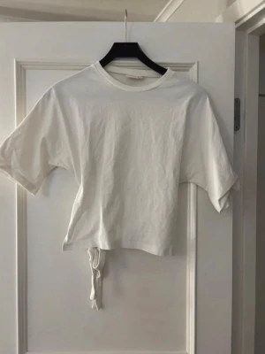 Vit croppad topp med knyt - Vit croppad topp i bomull med korta ärmar och snygg knytning i ryggen. Enkel och stilren design som passar perfekt till jeans eller kjol. T-shirten har rund halsringning och är superbekväm.